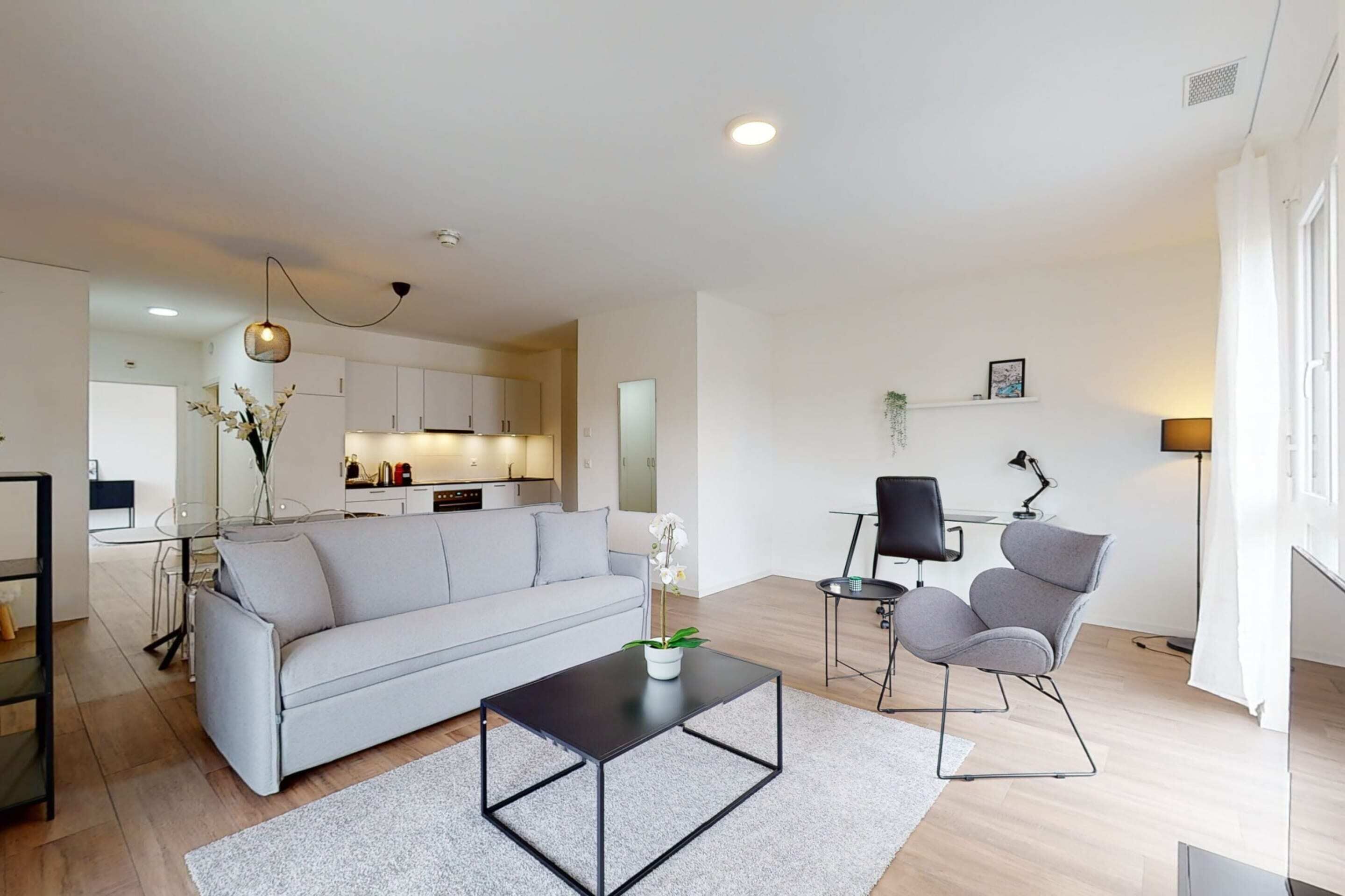 A modern living room with a grey sofa, a black coffee table, and a small side table with a lamp. A kitchen is visible in the background with white cabinets and a dining area.