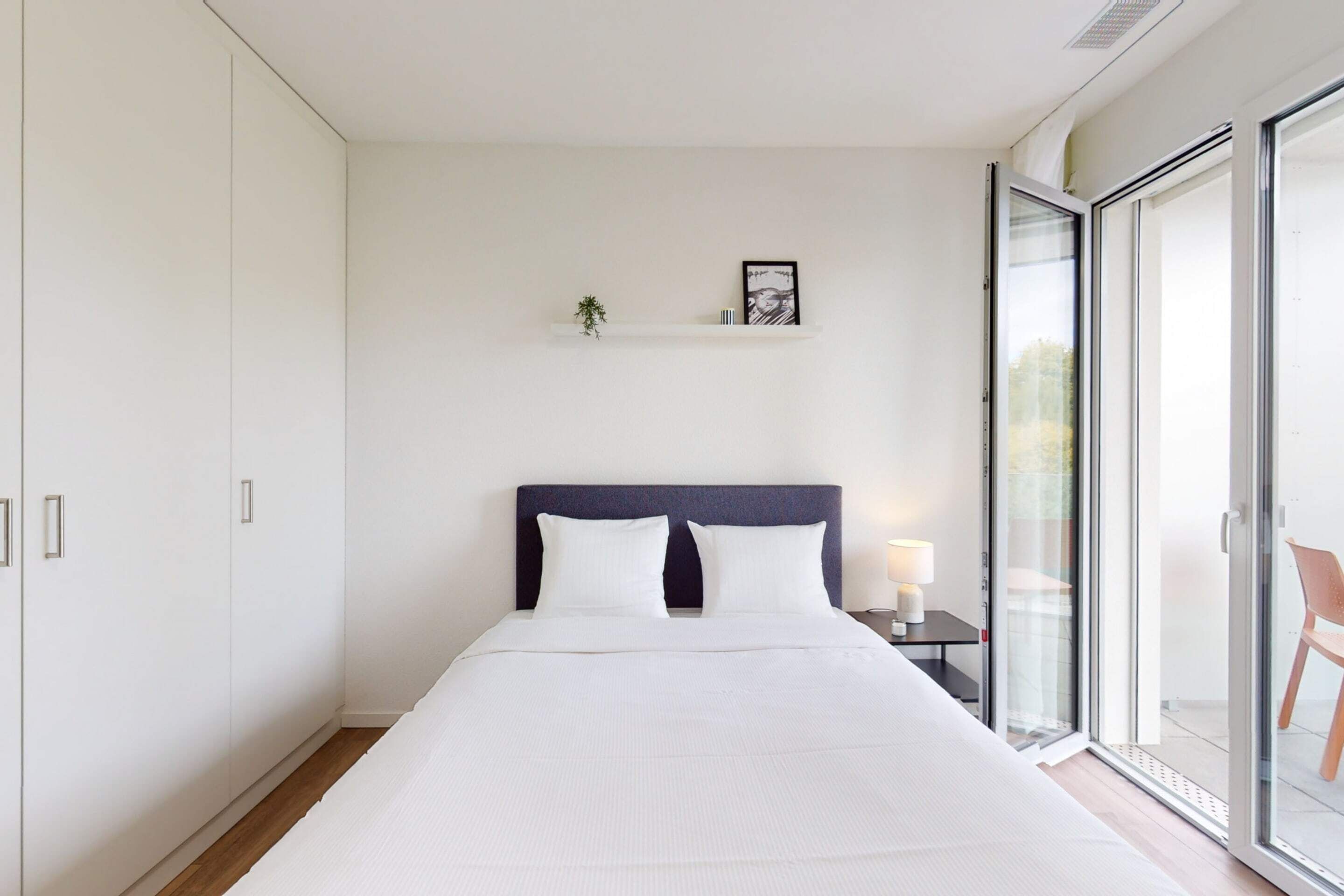 Un dormitorio moderno con una cama grande, un cabecero oscuro, una mesita de noche pequeña con una lámpara y una puerta corrediza de vidrio que da a un espacio exterior.