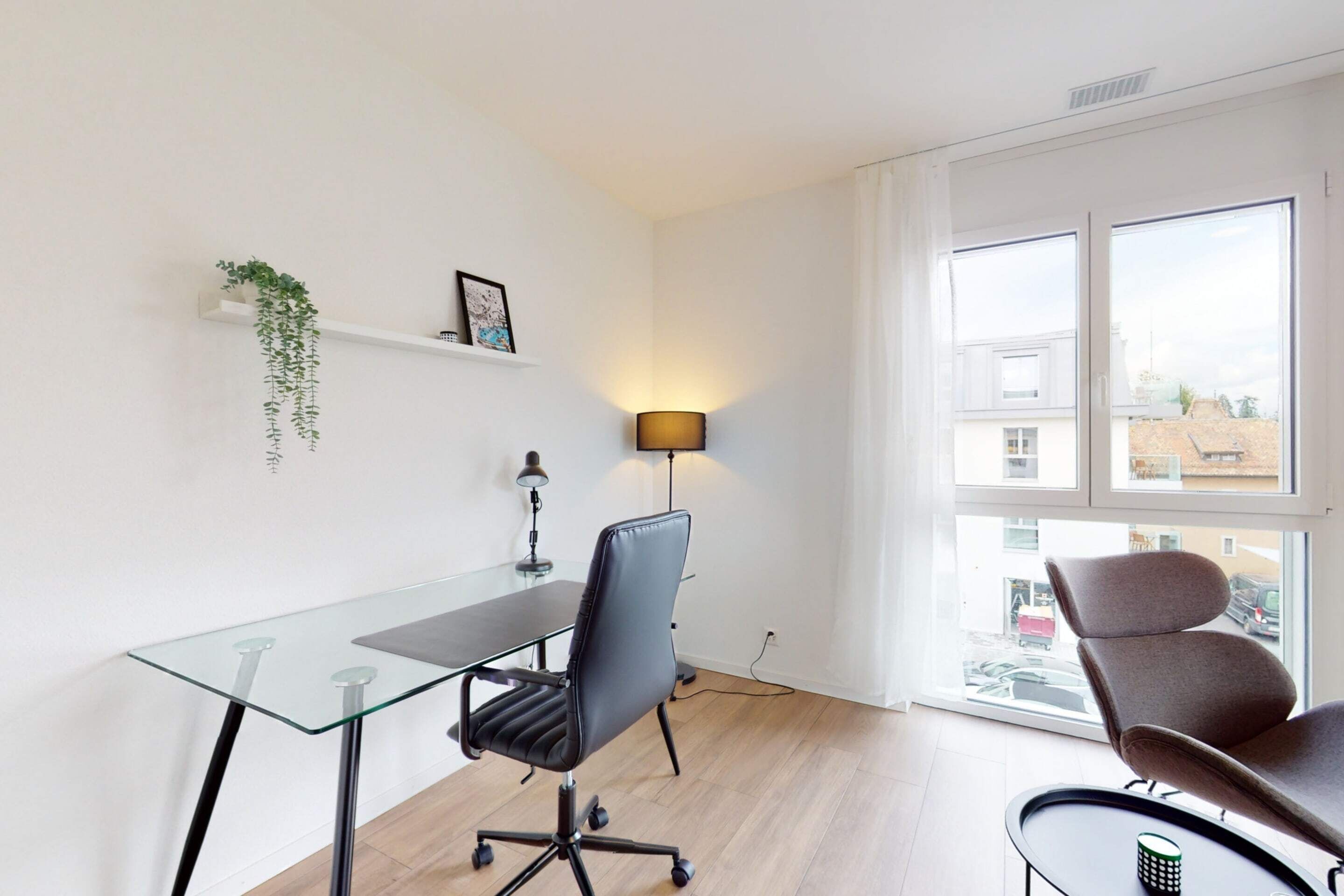 A modern office space with a glass desk, black office chair, and a lamp. There is a window with a view of the outside.