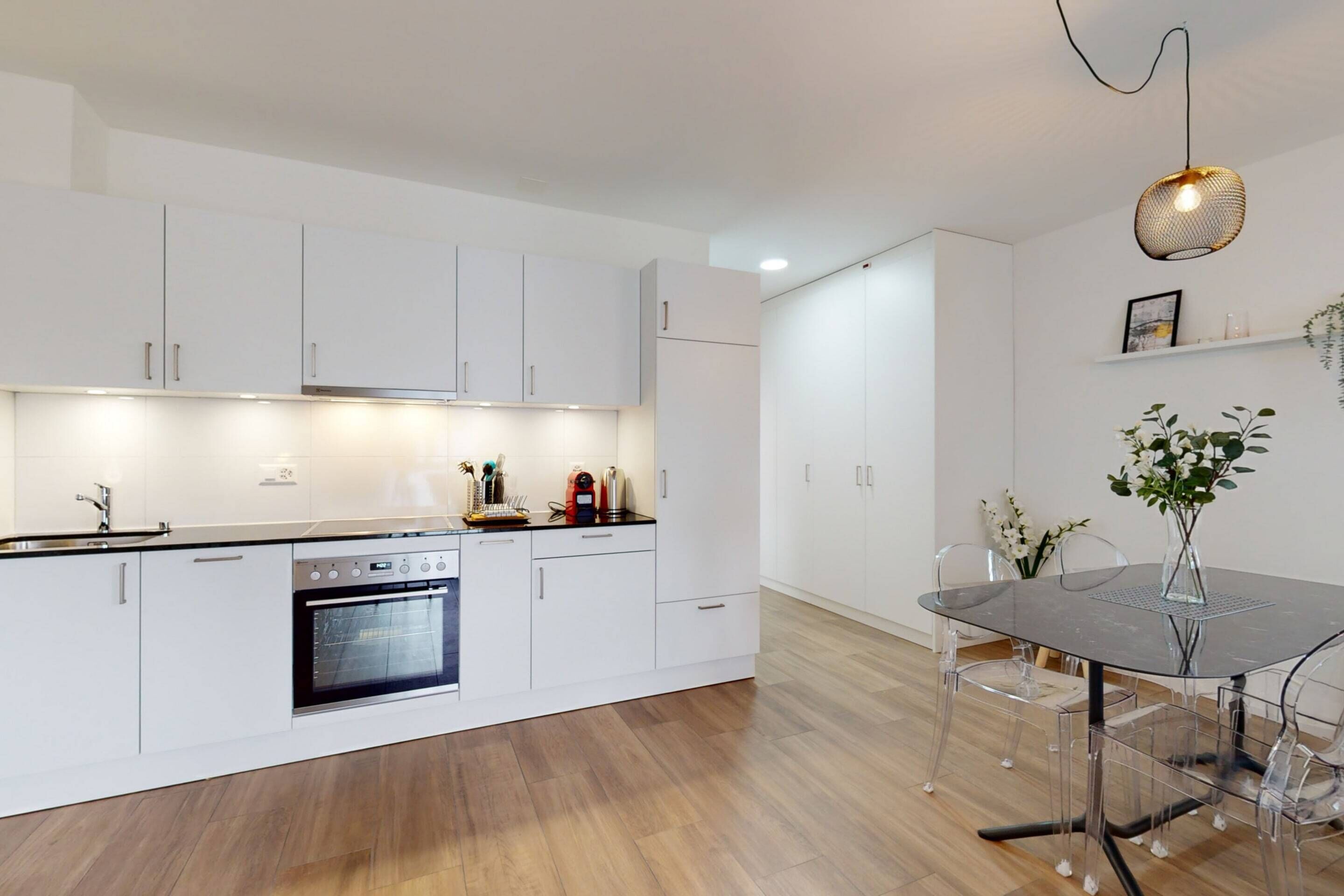 A modern kitchen with white cabinets, a built-in oven, and a dining area with a glass table and chairs.