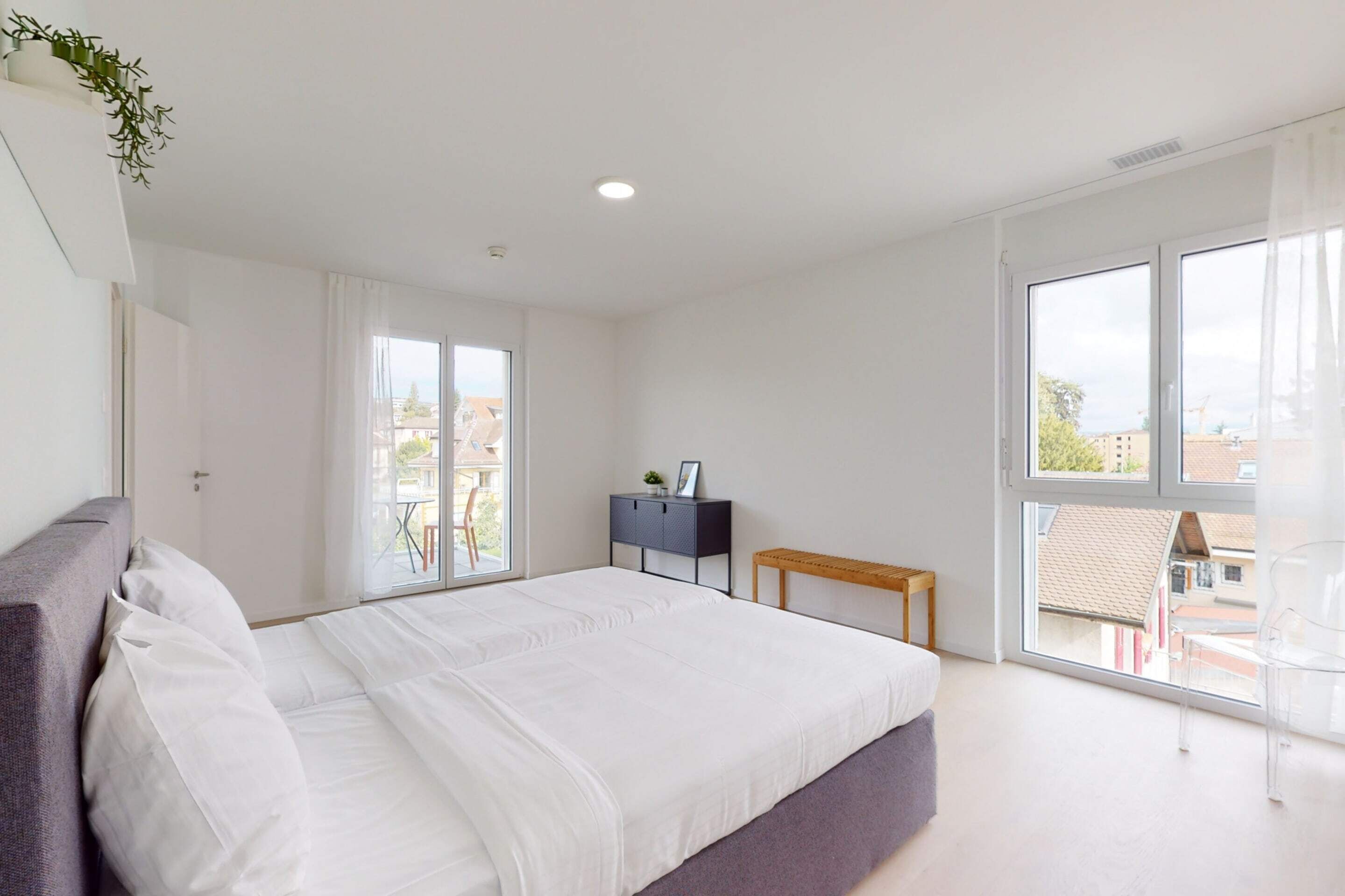Een moderne slaapkamer met een groot bed, een klein bureau en een raam met uitzicht op gebouwen.