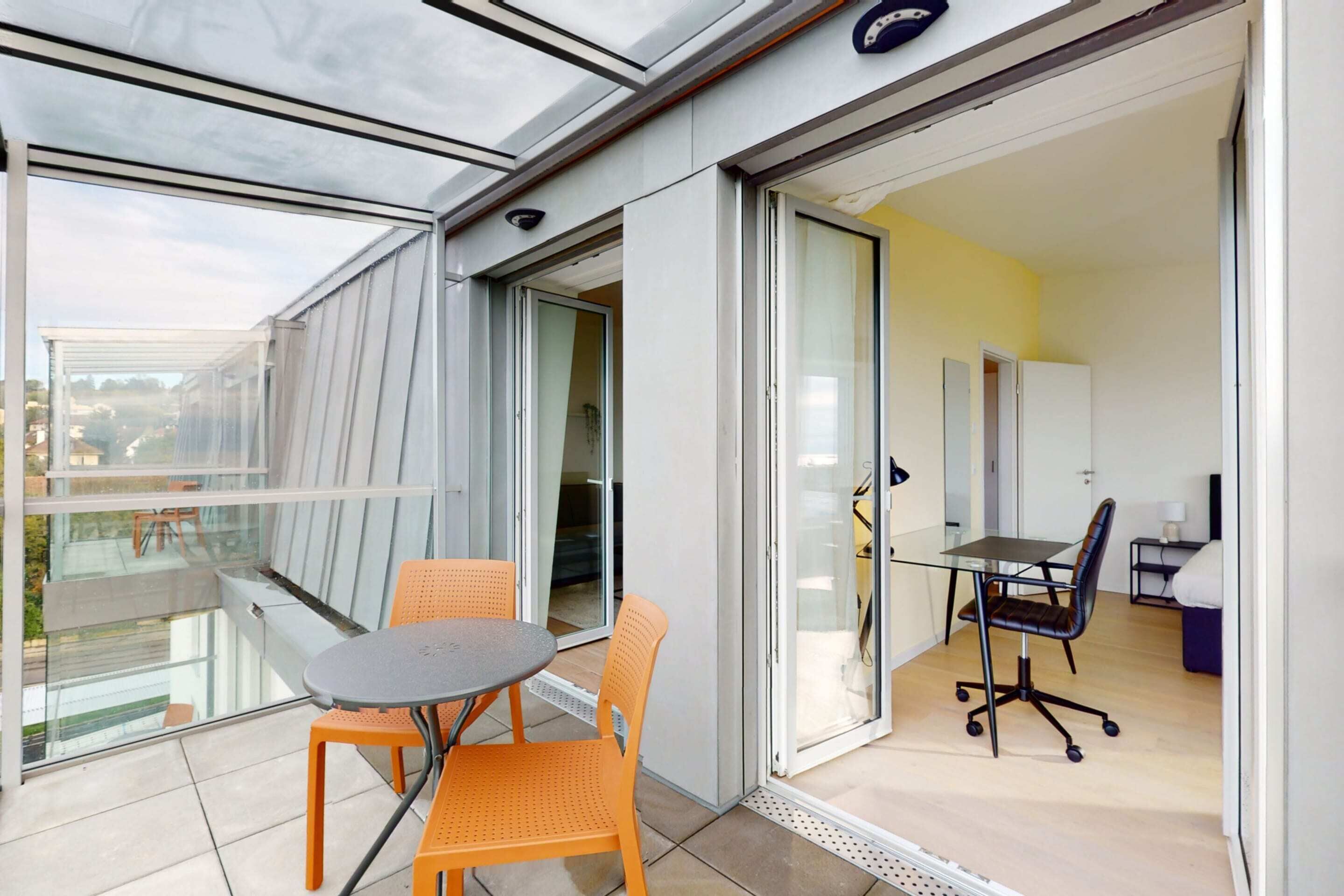 Un balcón con puerta de cristal que da a una habitación con una mesa y sillas, una silla con ruedas y vistas al exterior.
