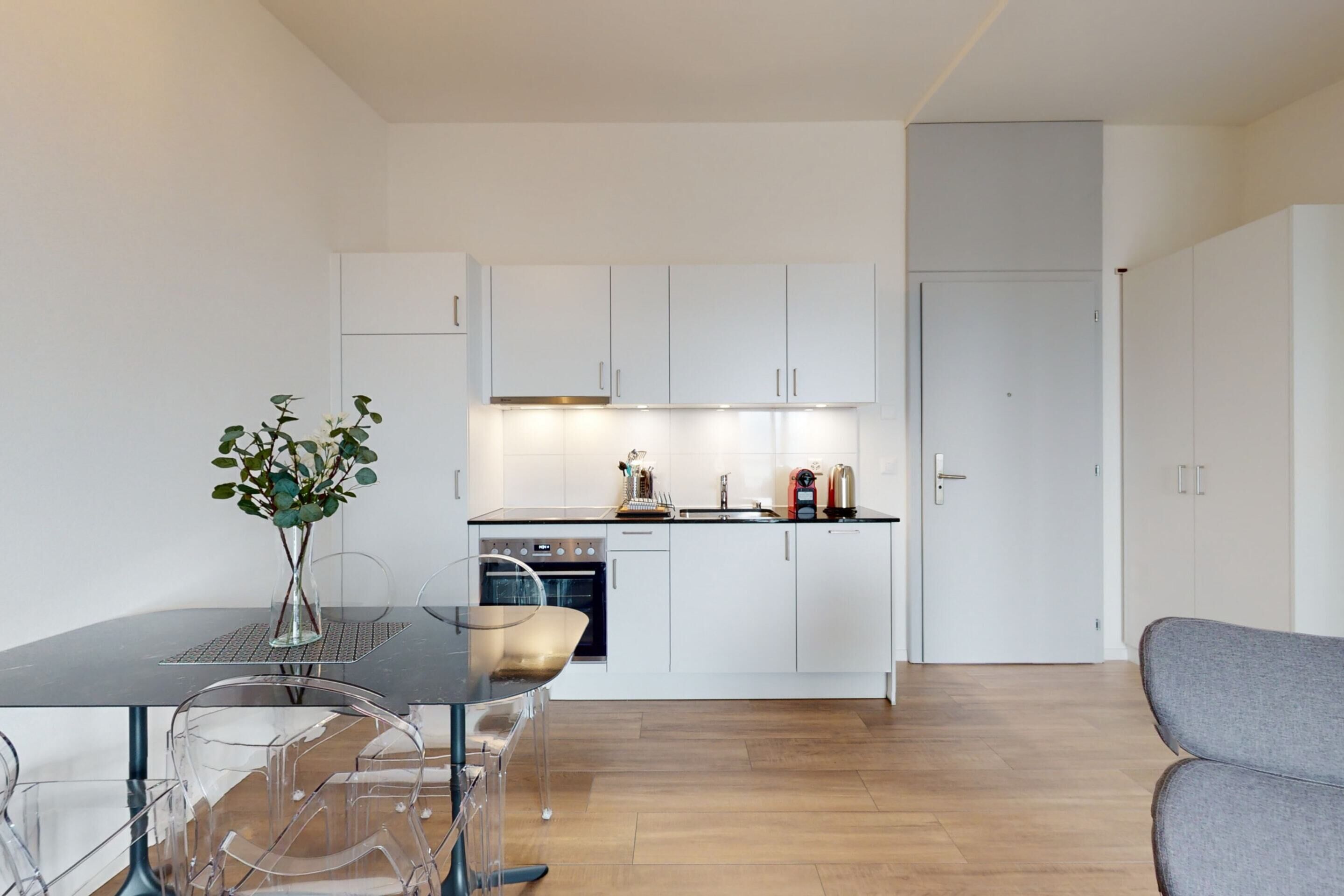 Una cocina moderna con gabinetes blancos, horno empotrado y comedor con mesa y sillas de vidrio.