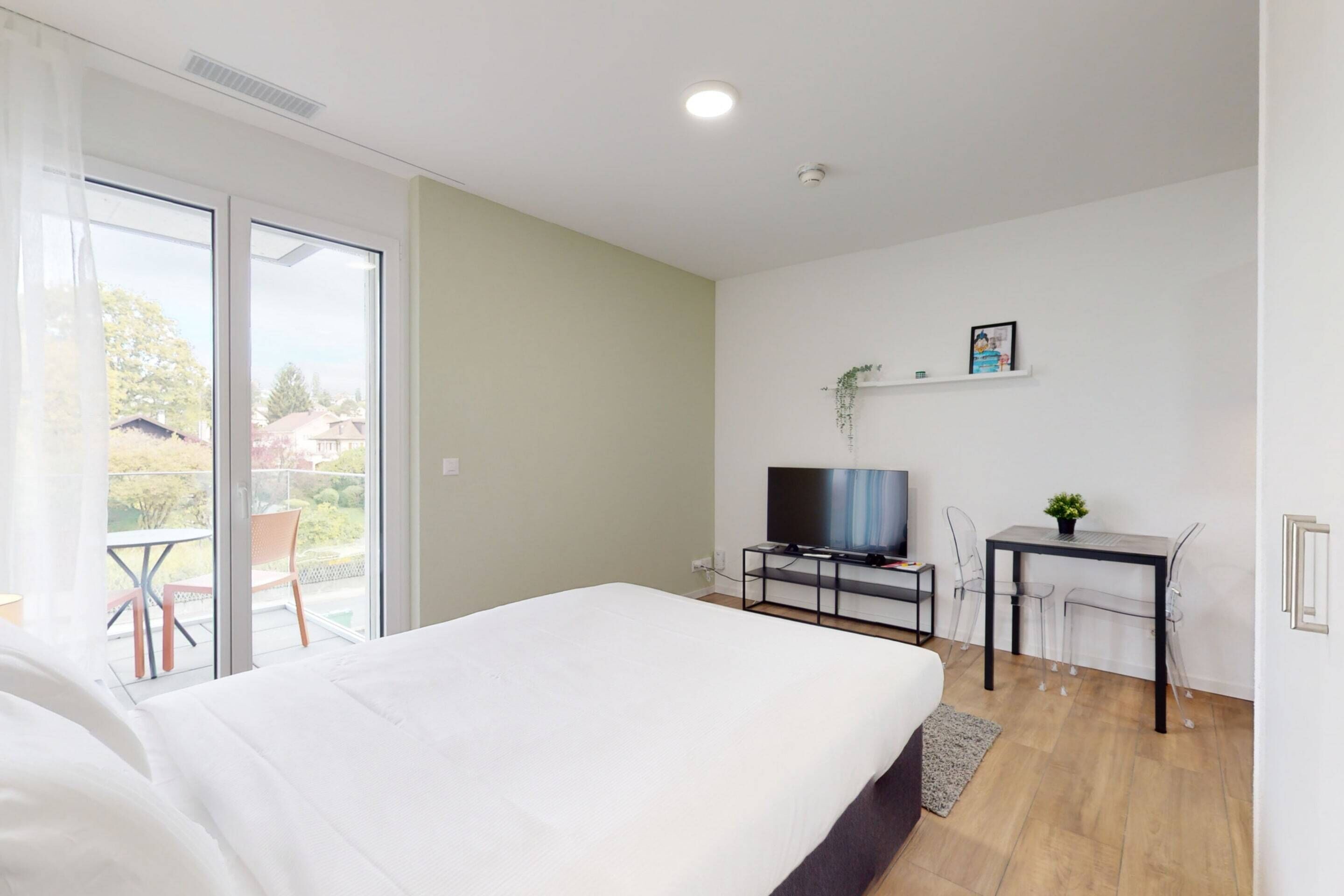 Un dormitorio moderno con una cama grande, un televisor en un mueble bajo, un escritorio pequeño con una silla y un ventanal corredero que da a un balcón.
