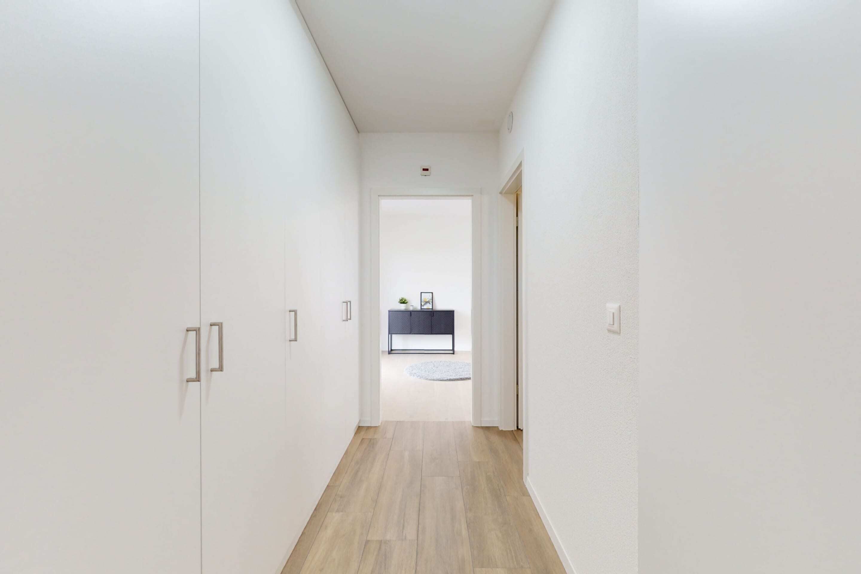 A narrow hallway with white walls and wooden flooring leading to a room with a dark-colored console table and a round rug.