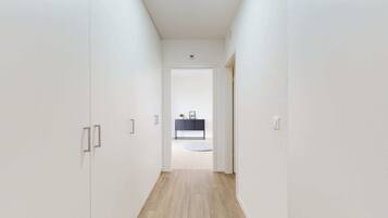 A narrow hallway with white walls and wooden flooring leading to a room with a dark-colored console table and a round rug.