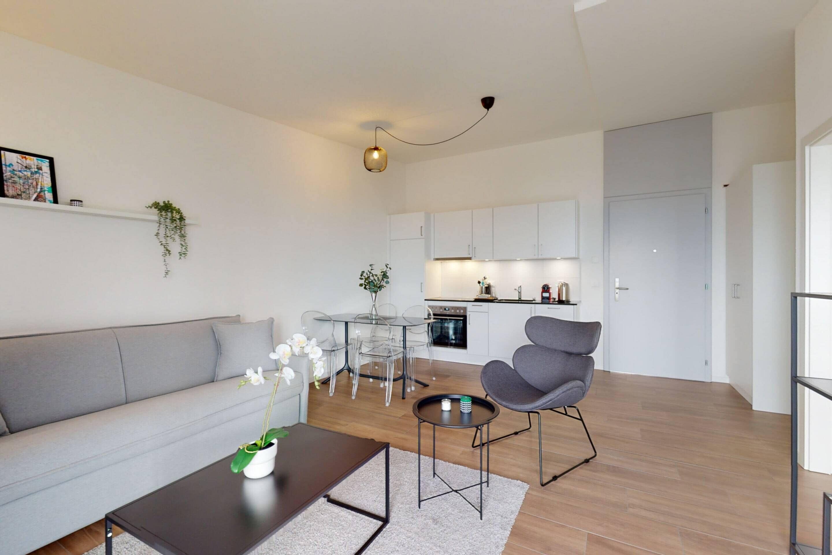 A modern living room with a grey sofa, a coffee table, and a grey armchair. There is a dining table with chairs and a kitchen area in the background.