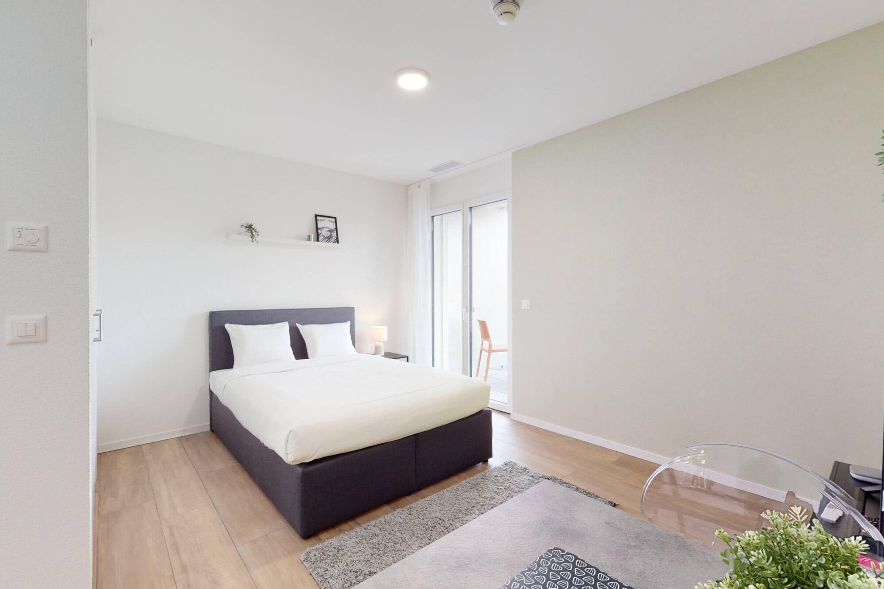 A modern bedroom with a large bed, a small bedside table, a chair, and a glass table.