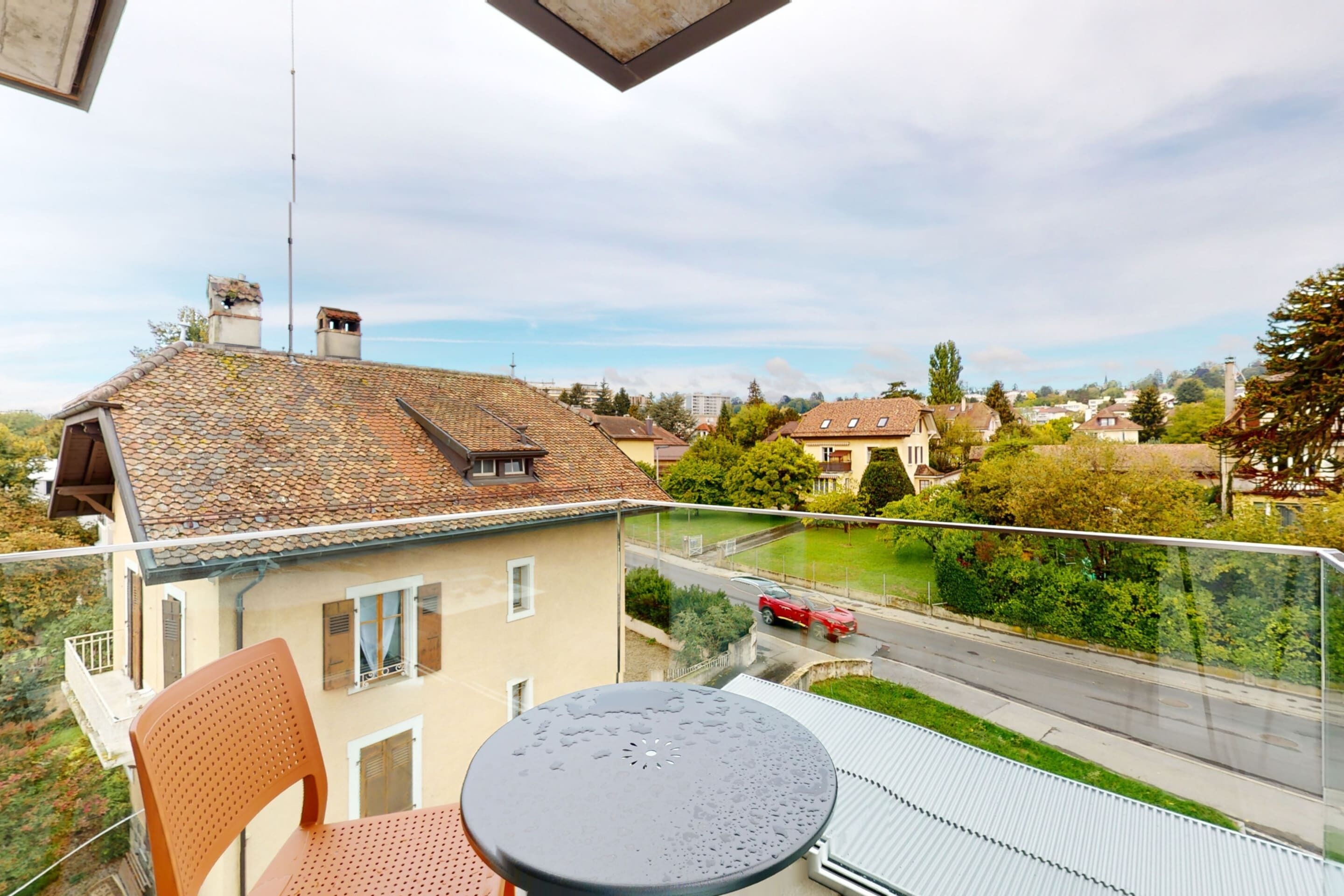 Un balcón con mesa y sillas, con vistas a una calle con casas y vegetación.