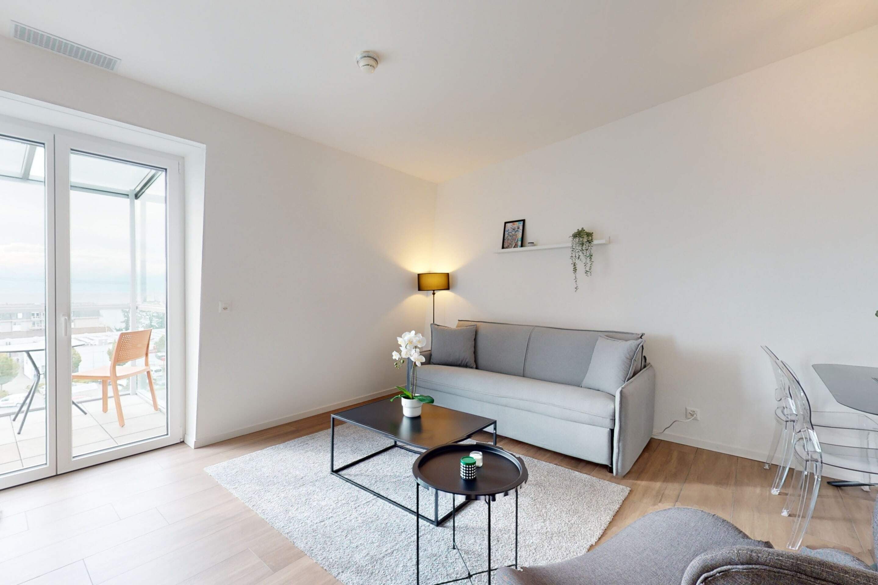Un salon moderne avec un canapé, une table basse et une lampe. On aperçoit un balcon avec une chaise à travers les portes vitrées.