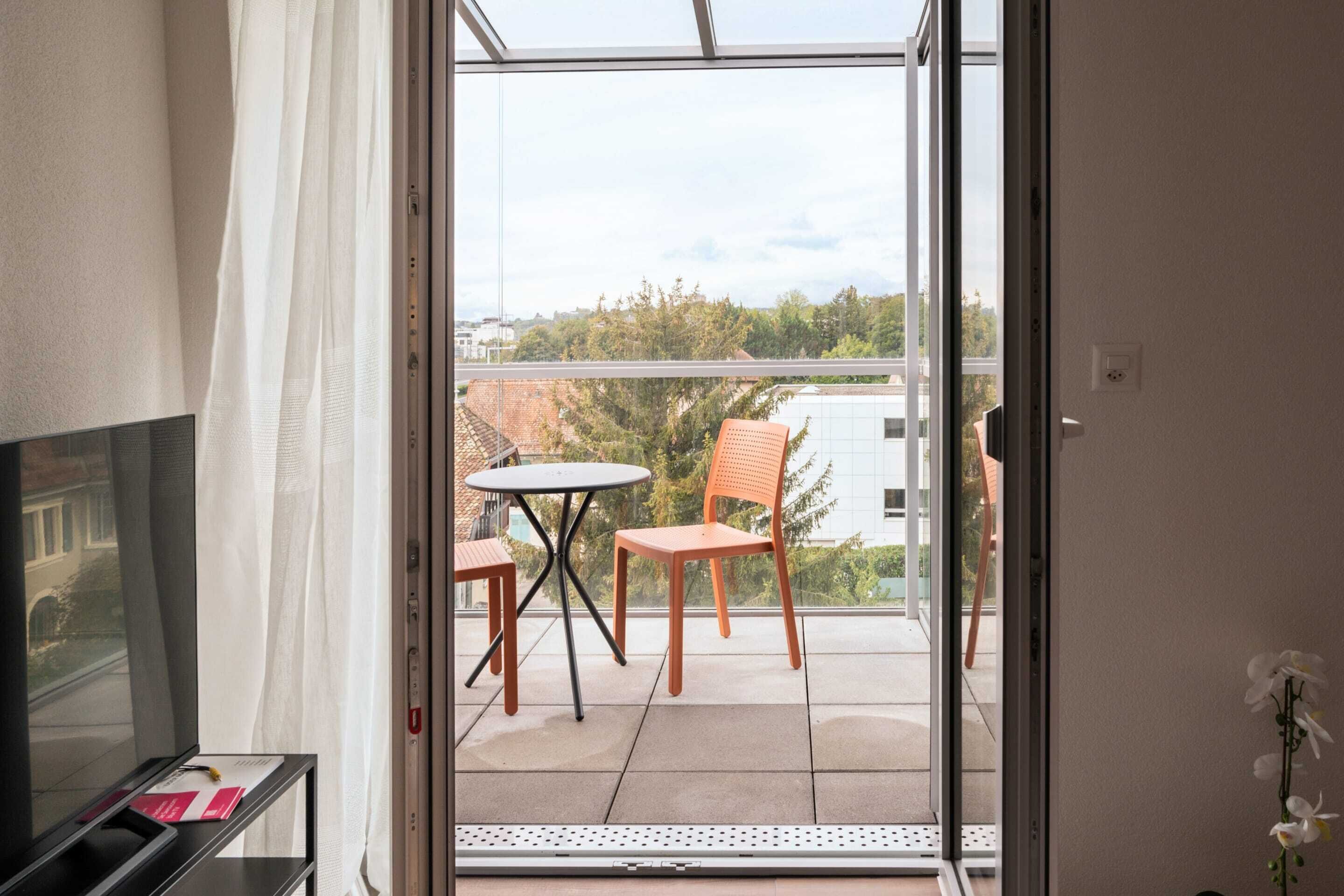 A balcony with a table and two chairs, a TV on a stand, and a view of buildings and trees.