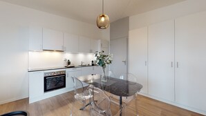 A modern kitchen with white cabinets, a black countertop, a glass dining table with clear chairs, and a hanging light fixture. - Lodges Hotel Morges (Morges)