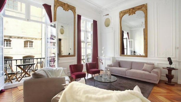 Amplia sala de estar con sofá color beige, sillones rojos y una mesa de centro de vidrio. Se observan espejos grandes y un balcón con mobiliario exterior.
