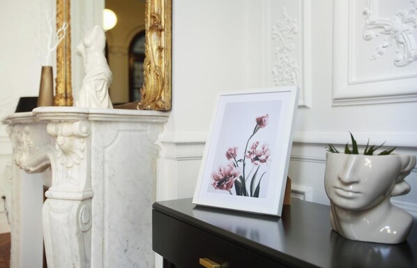 Une photo encadrée de fleurs roses sur un fond noir, un vase blanc orné d’un motif représentant un visage et un miroir avec un cadre doré.