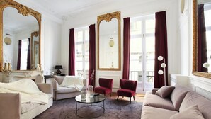 Un salón amplio con un sofá color beige, un sillón rojo y una mesa de centro redonda. Se observan espejos grandes y cortinas rojas.