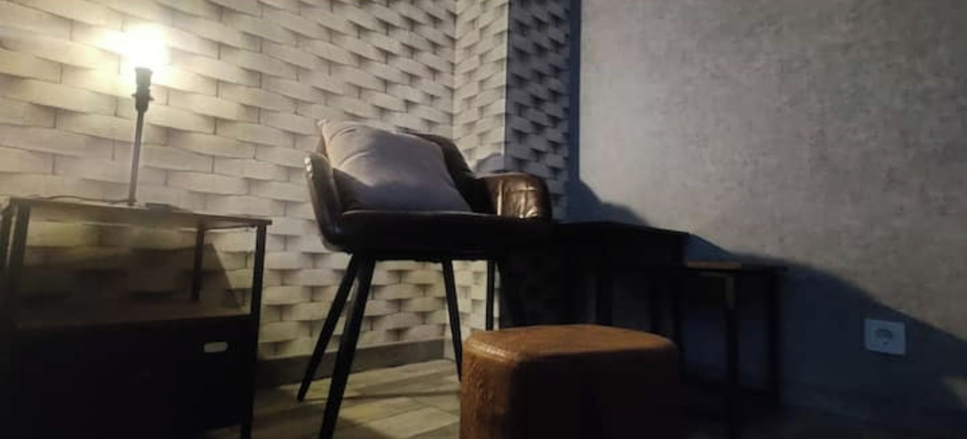 A room with a brick-patterned wall, a black chair, a small wooden stool, and a table with a glass top.