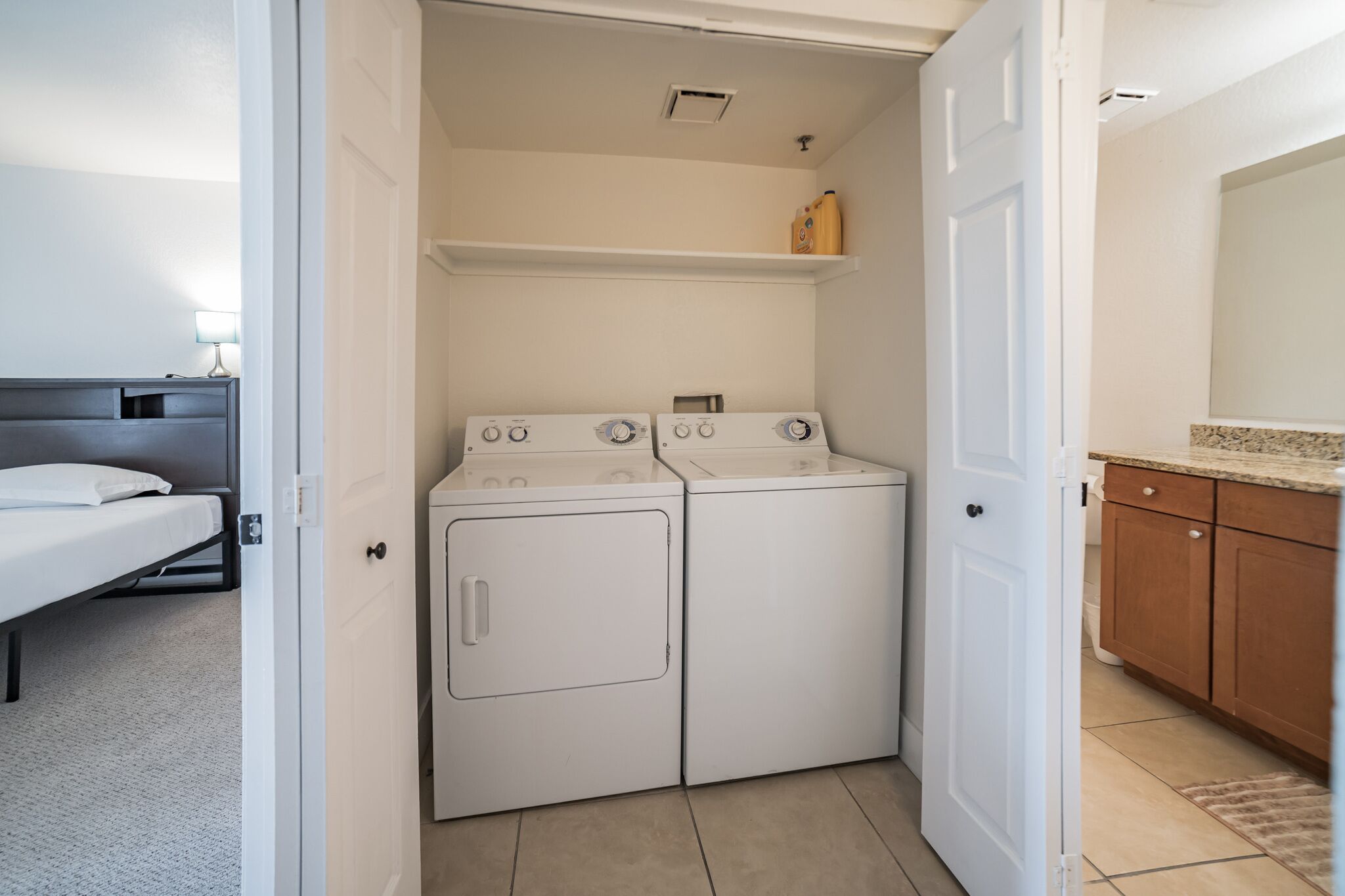 A compact laundry room with a washer and dryer, a bed in the adjacent room, and a small shelf with cleaning supplies.