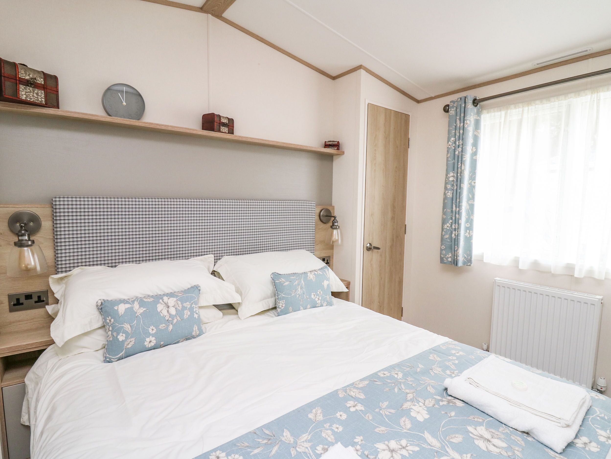 A bedroom with a bed, a nightstand, a wall clock, a shelf with decorative items, and a window with curtains.
