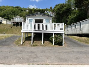 A single-story mobile home with a balcony, surrounded by a grassy area and trees. - Balmoral (Carmarthen)