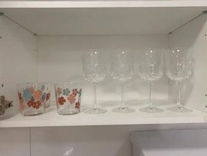 Uma prateleira branca com quatro taças de vinho transparentes e dois recipientes de vidro com desenhos florais.