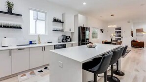 A modern kitchen with white cabinetry, a central island with bar stools, and a dining area with a sofa and table. - Modern Luxury In The City at Havilah Estates (Atlanta)