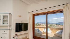 A room with a TV mounted on the wall, a cabinet with drawers, a sliding glass door leading to an outdoor seating area with white furniture, and a view of the landscape. - Casa Lola by Wonderful Italy (Olbia)