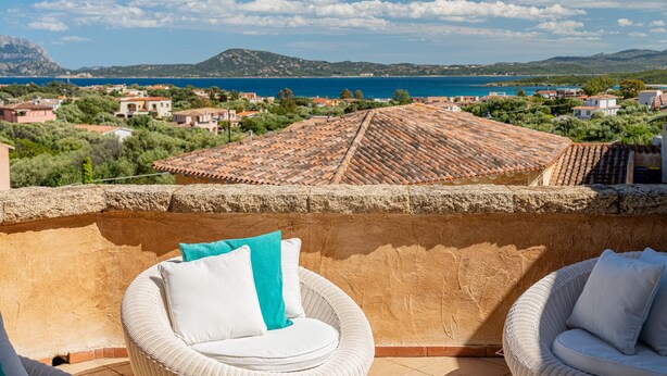 A balcony with wicker chairs and cushions, overlooking a coastal town with terracotta roofs and a clear blue sea. - Casa Lola by Wonderful Italy (Olbia)