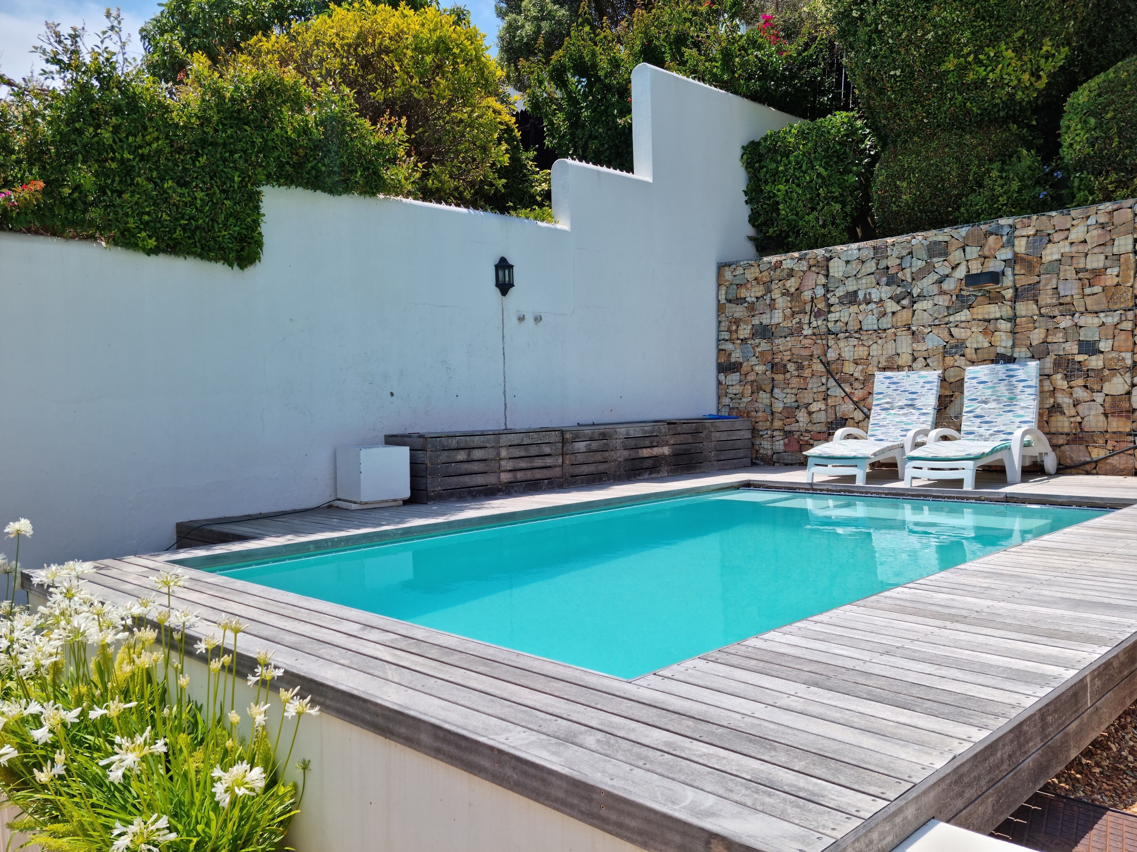 A swimming pool with a wooden deck, white walls, and a stone wall with a stone mosaic. There are two white lounge chairs and a small white heater.