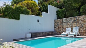 Una piscina amb un pont de fusta, parets blanques i una paret de pedra amb una mosaic de pedra. Hi ha dos gandules blanques i un petit escalfador blanc.