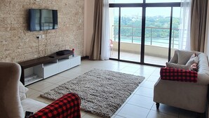 Ein geräumiges Wohnzimmer mit großem Fenster, Deckenventilator, Flachbildfernseher an einer Ziegelwand und zwei Sofas mit rot-schwarz kariertem Überwurf.