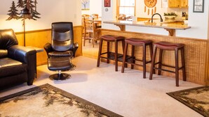 Een ruime woonkamer met een plafondventilator, een leren fauteuil, een eettafel en een keuken op de achtergrond.