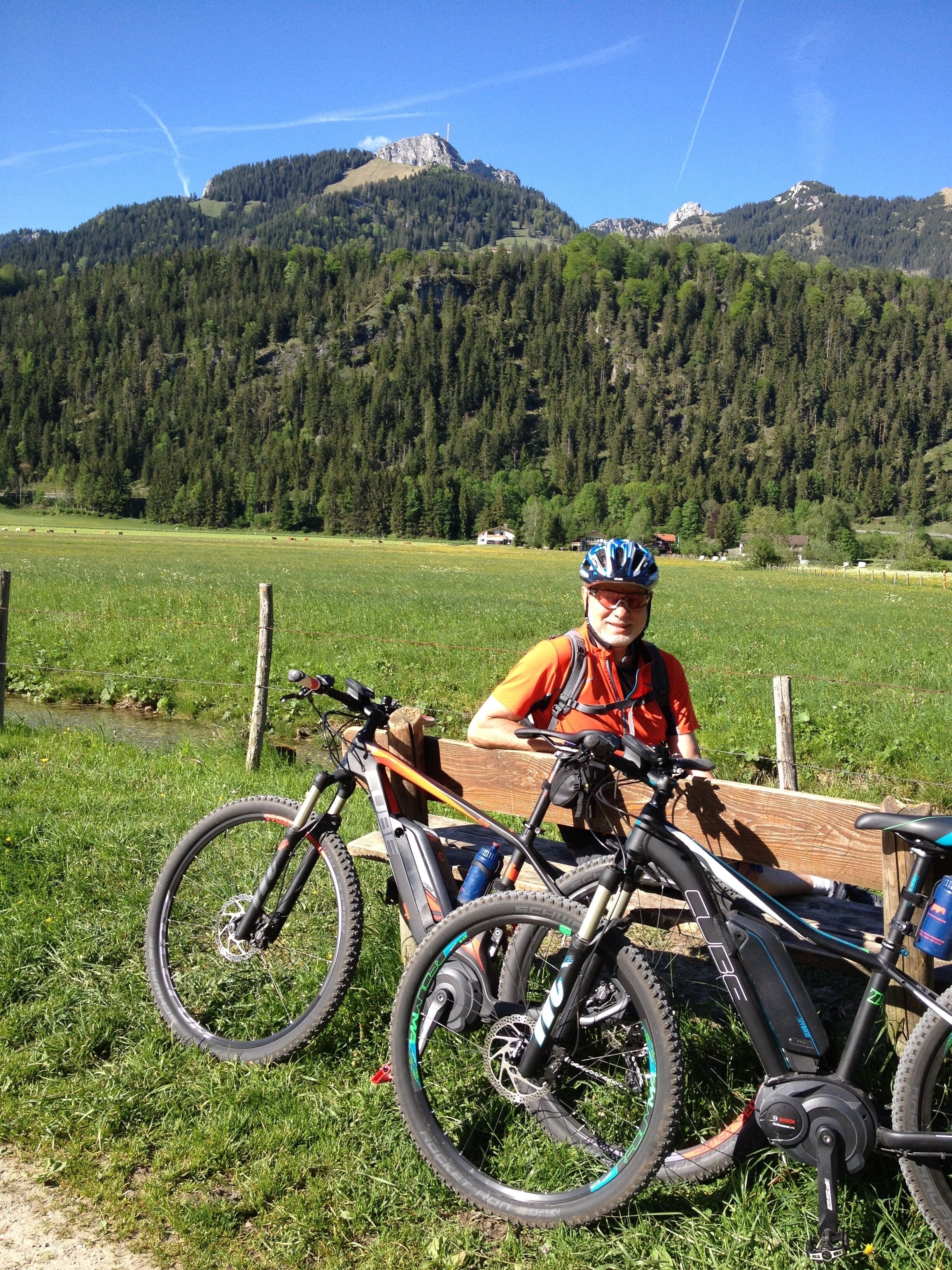 Una persona sentada en un banco en un campo cubierto de hierba, con dos bicicletas y montañas al fondo.