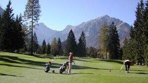 Un parcours de golf avec une chaîne de montagnes en arrière-plan, un chariot et un golfeur qui se prépare à frapper la balle.