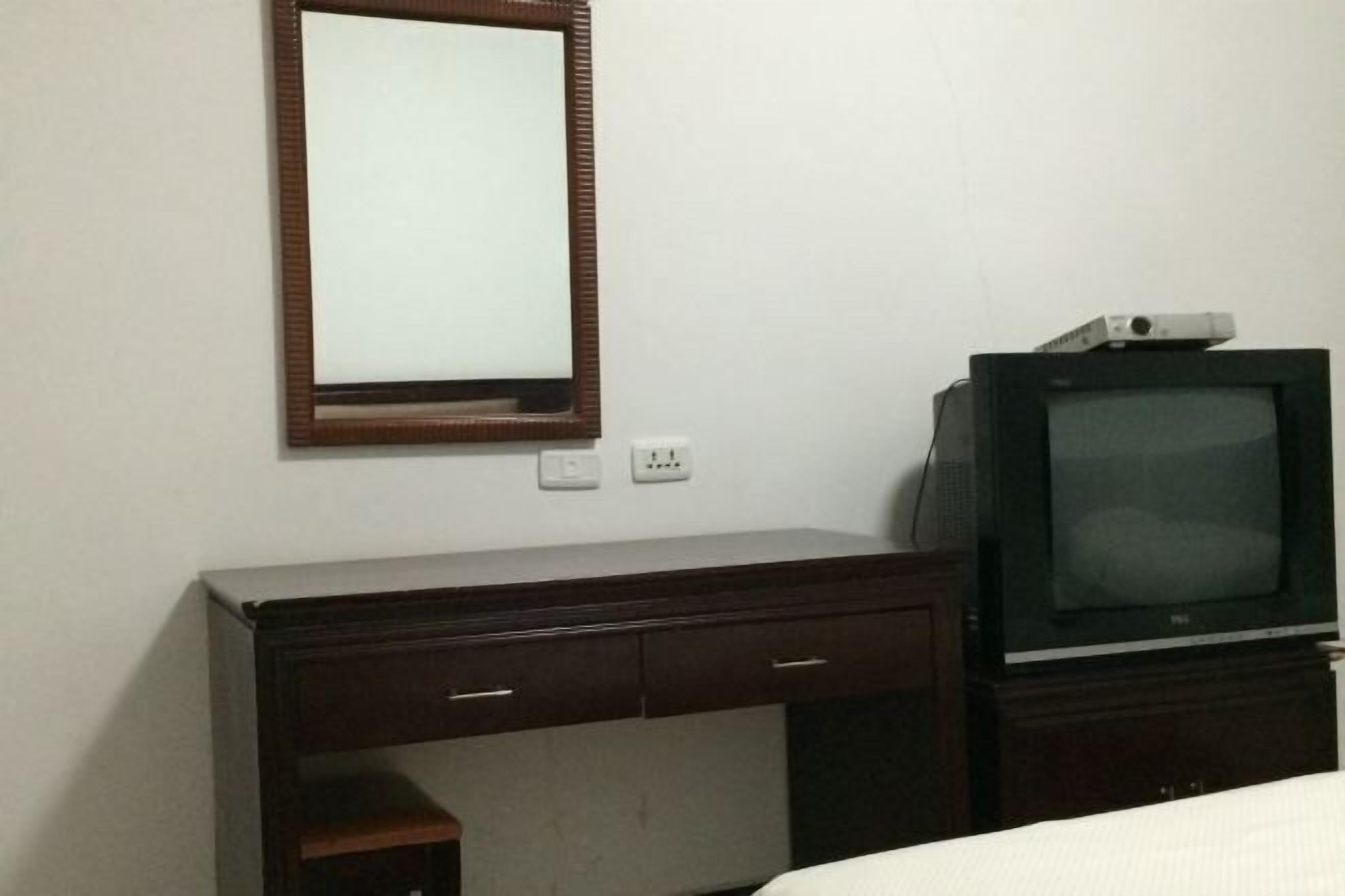 Quarto com escrivaninha de madeira, televisão em um suporte e espelho na parede.