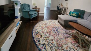 Ein modernes Wohnzimmer mit Fernseher, einem smaragdgrünen Sessel, einem grauen Sofa und einem farbenfrohen Teppich.