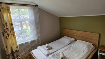 Un lit simple avec du linge de lit blanc et deux serviettes enroulées, une tête de lit en bois, une fenêtre donnant sur des immeubles et un luminaire au plafond.