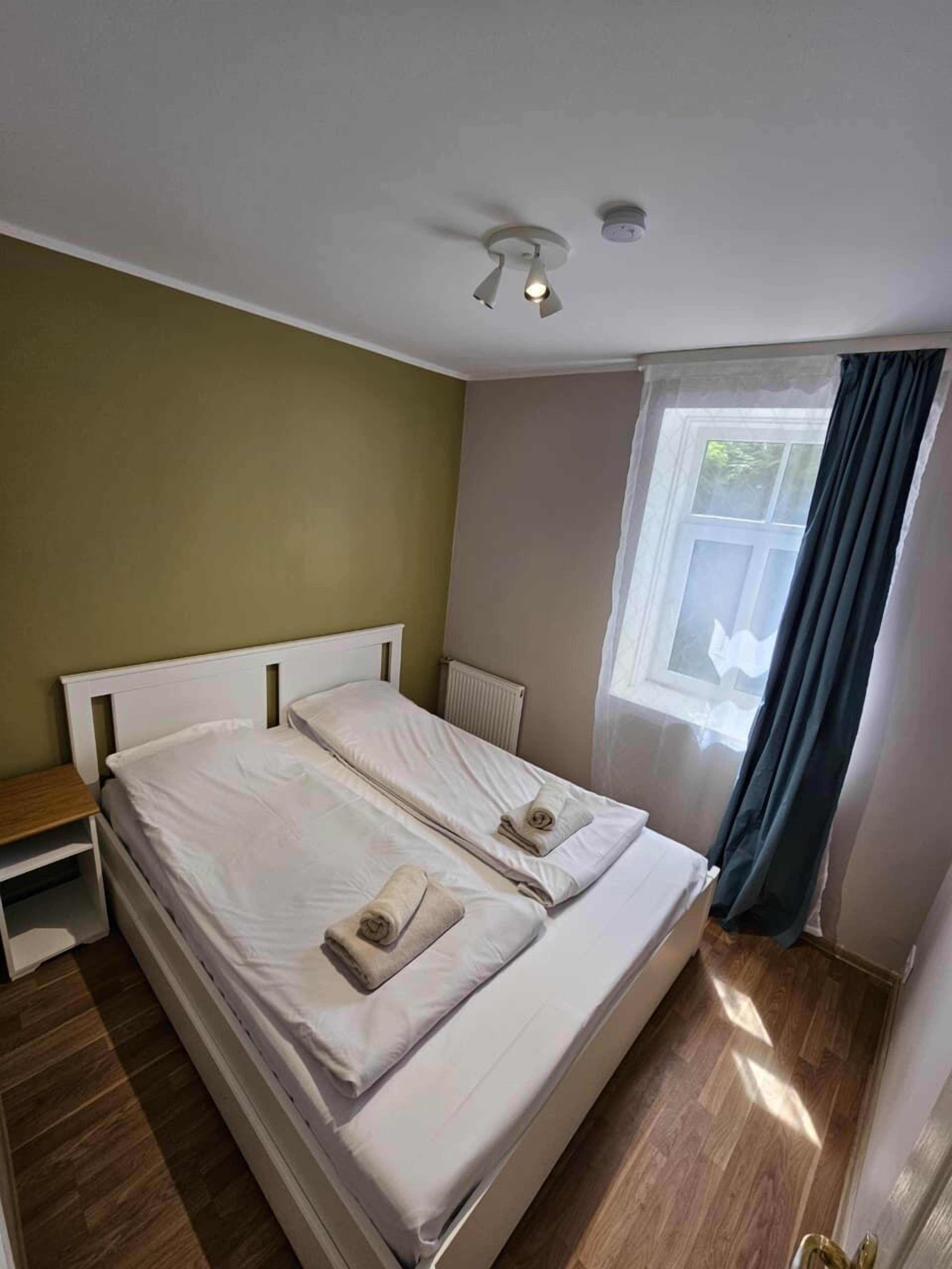 Una cama doble con sábanas y toallas blancas, una mesita de noche, una ventana con cortinas y un ventilador de techo.