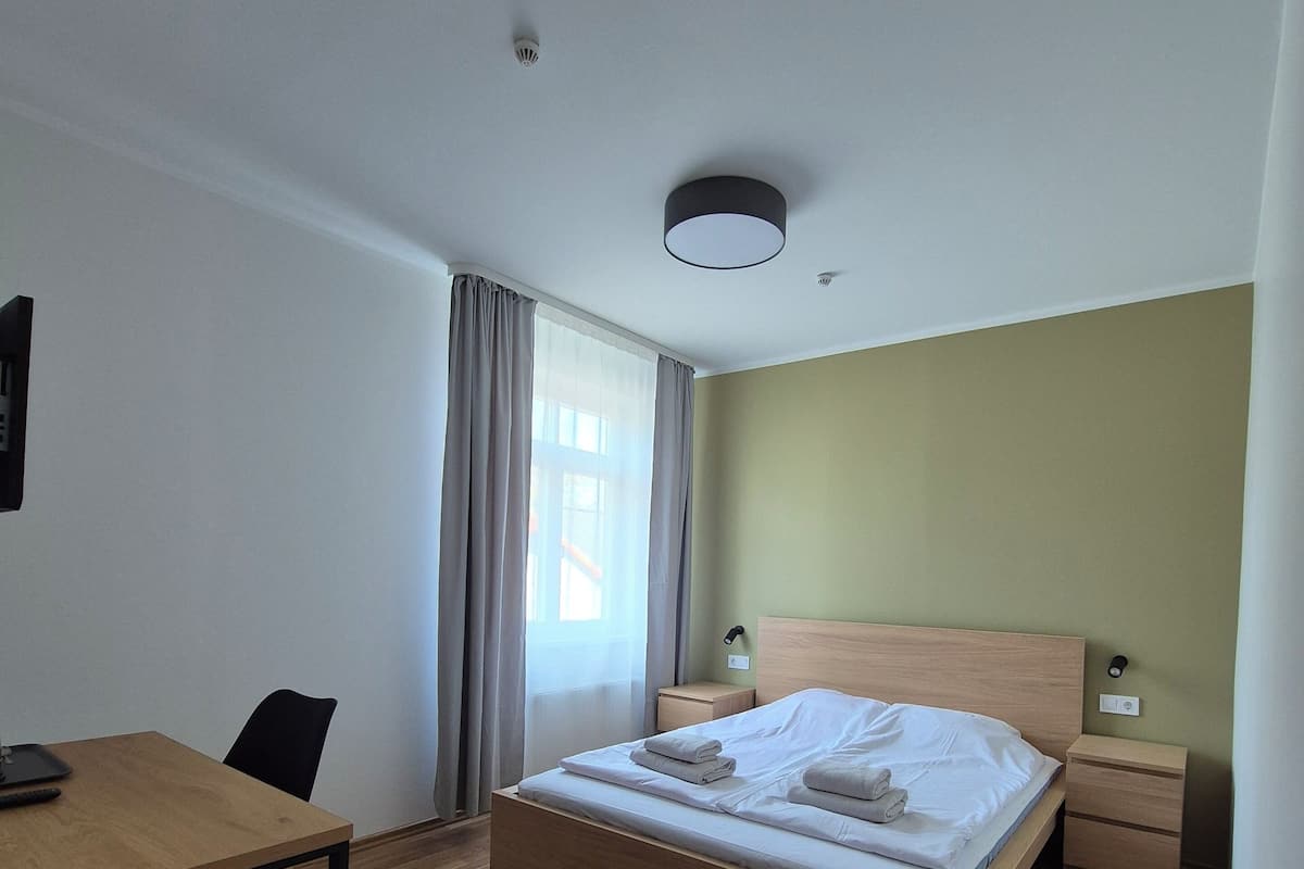 A hotel room with a wooden bed, a desk with a chair, a nightstand, and a wall-mounted lamp.