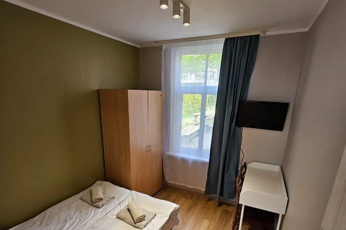 A small, single-bed room with a wooden wardrobe, a bedside table, and a window with curtains.