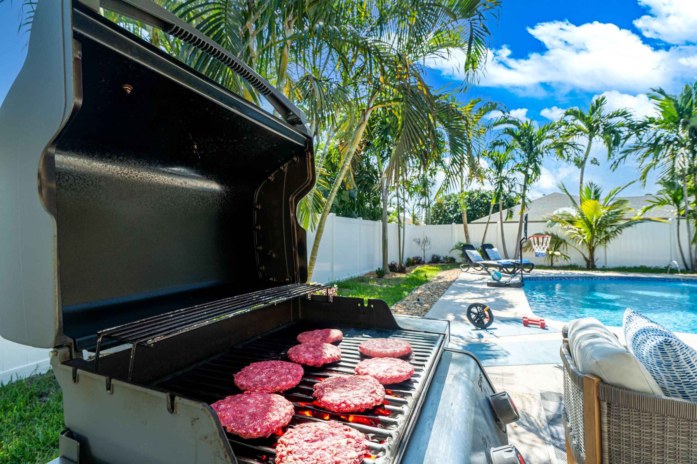 A backyard with a grill cooking hamburgers, a pool, and lounge chairs.