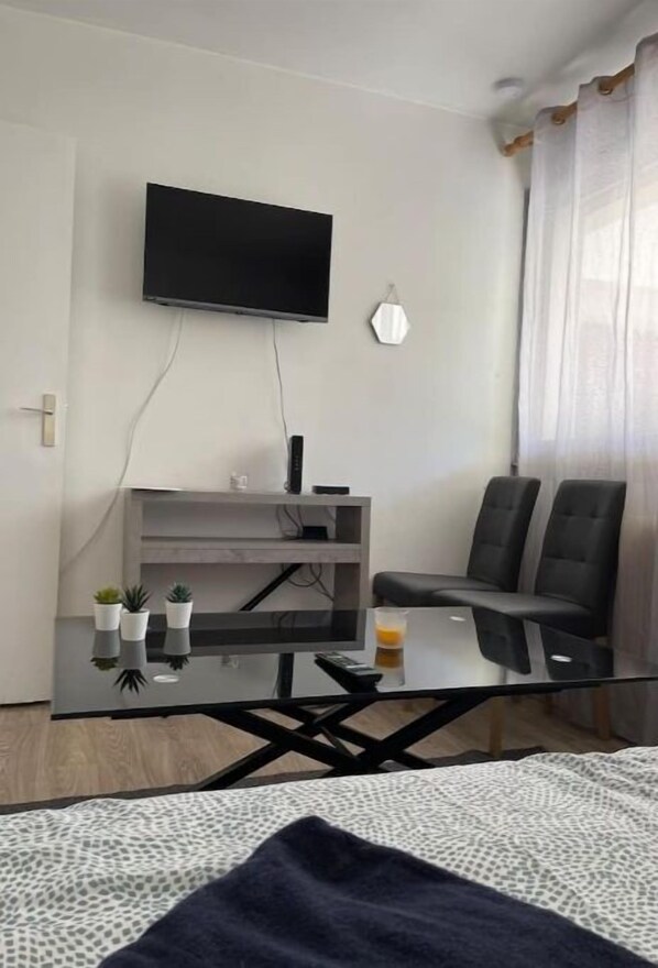 Ein modernes Zimmer mit einem wandmontierten Flachbildfernseher, einem Glastisch, zwei schwarzen Stühlen, einer kleinen Ablage mit Pflanzen und einem Bett mit einem gemusterten Überwurf.