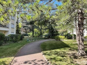 Ein geschwungener Weg in einer begrünten Anlage mit Bäumen und einem Gebäude im Hintergrund.