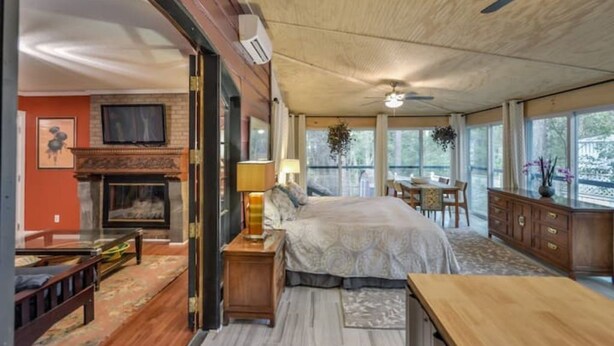 A spacious bedroom with a large bed, a fireplace, a dining area with a table and chairs, and a view through large windows. - Eclectic Forest View Efficiency in Quiet Chapel Hill Neighborhood (Chapel Hill)