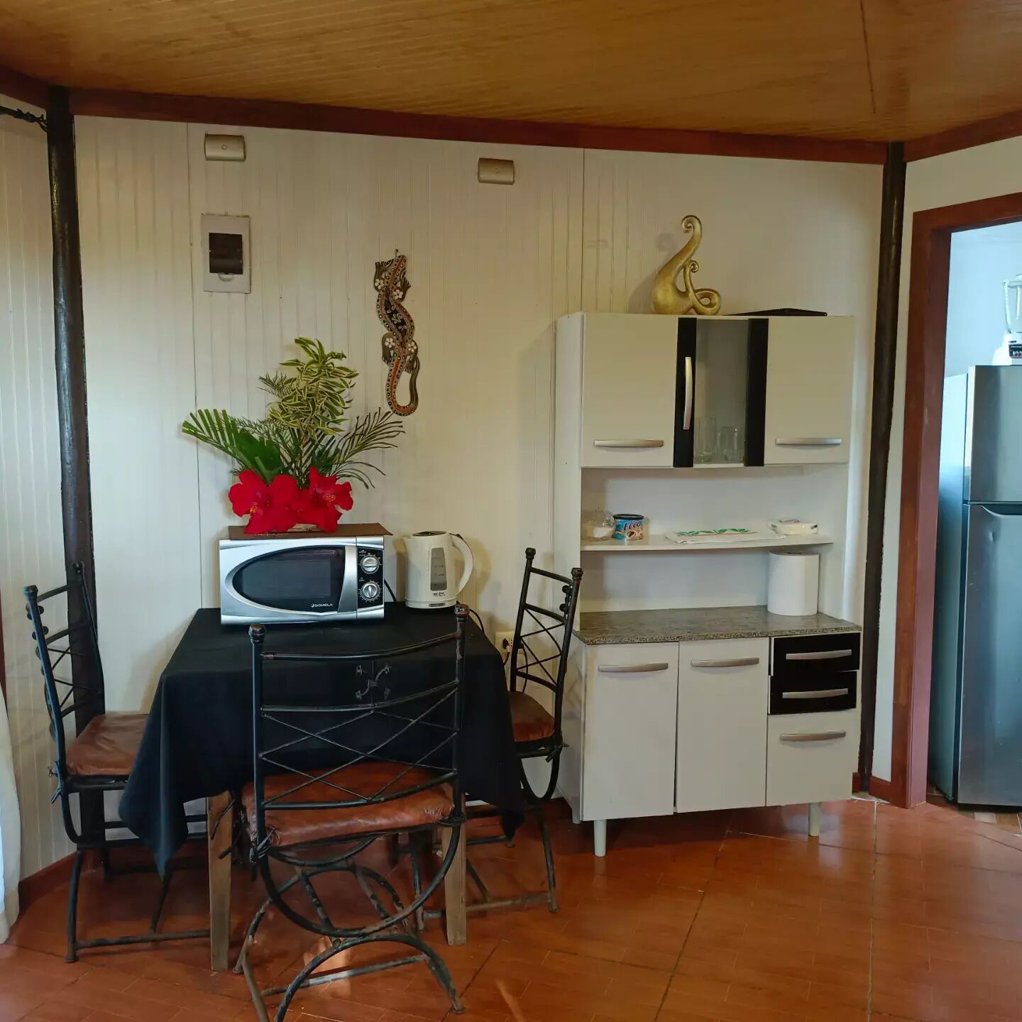 A compact kitchen with a microwave, kettle, and small cabinet. There is a dining table with chairs and a potted plant.