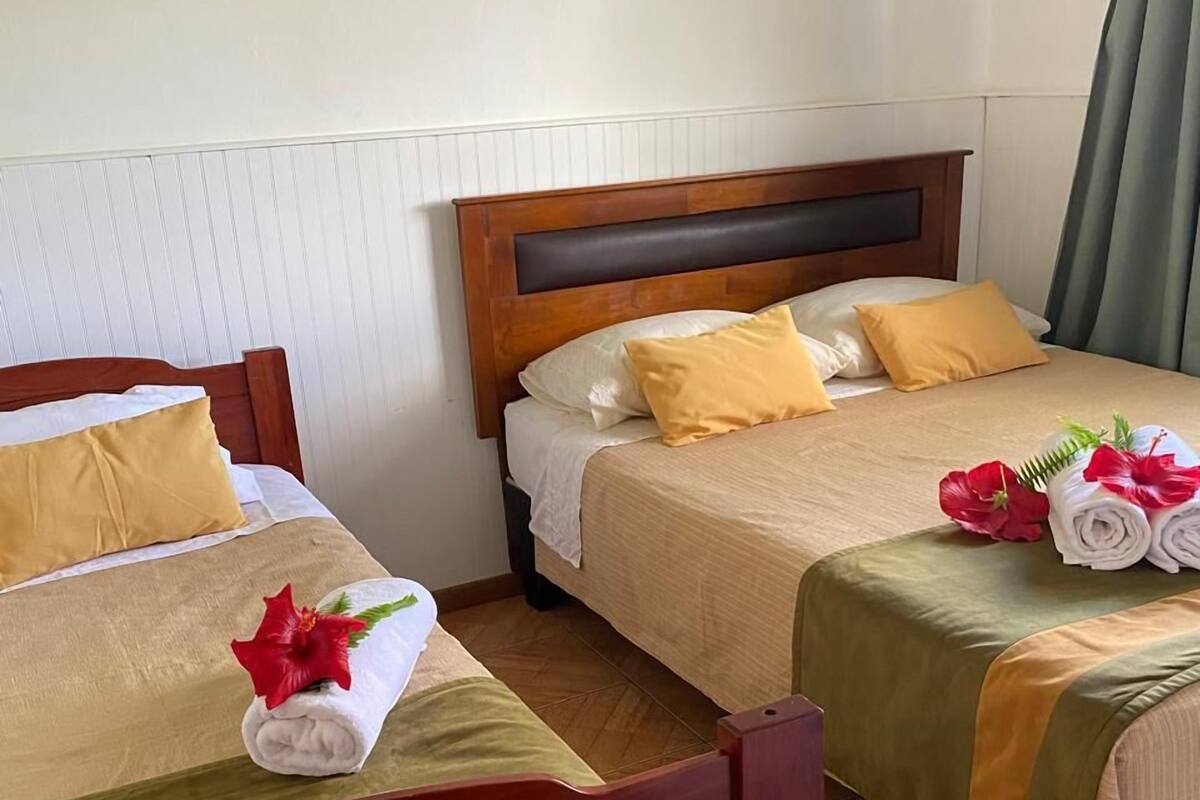 A hotel room with two wooden beds, each with a headboard, a green and beige bedspread, and a yellow pillow. There is a wall-mounted artwork of a red flower.