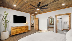 A bedroom with a large bed, wooden ceiling, ceiling fan, TV, and a potted plant. - Moonlight Lodge |  Private Hot Tub | Zion NP (Orderville)