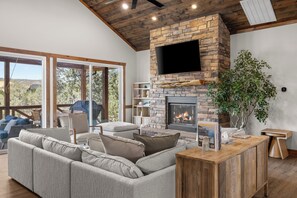 A modern living room with a stone fireplace, a large flat-screen TV, and a view of the outdoors.