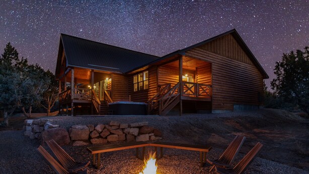 A log cabin with a fire pit and two lounge chairs, surrounded by a stone wall and a gravel area. - Moonlight Lodge |  Private Hot Tub | Zion NP (Orderville)