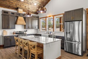A modern kitchen with dark cabinetry, a stainless steel refrigerator, and a central island with bar stools. - Moonlight Lodge | Private Hot Tub | Zion NP (Orderville)