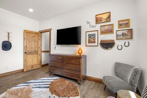 A room with a wooden door, a flat-screen TV mounted on the wall, a wooden dresser, a patterned rug, and a chair with a checkered cushion. - Moonlight Lodge | Private Hot Tub | Zion NP (Orderville)